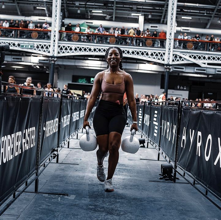 A female fitness competitor executing a heavy farmers carry walk with kettlebells during a HYROX event in Singapore.