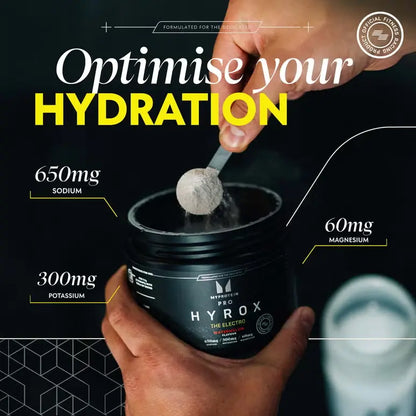 Scoop of Myprotein HYROX THE Electro powder above an open tub beside a water bottle
